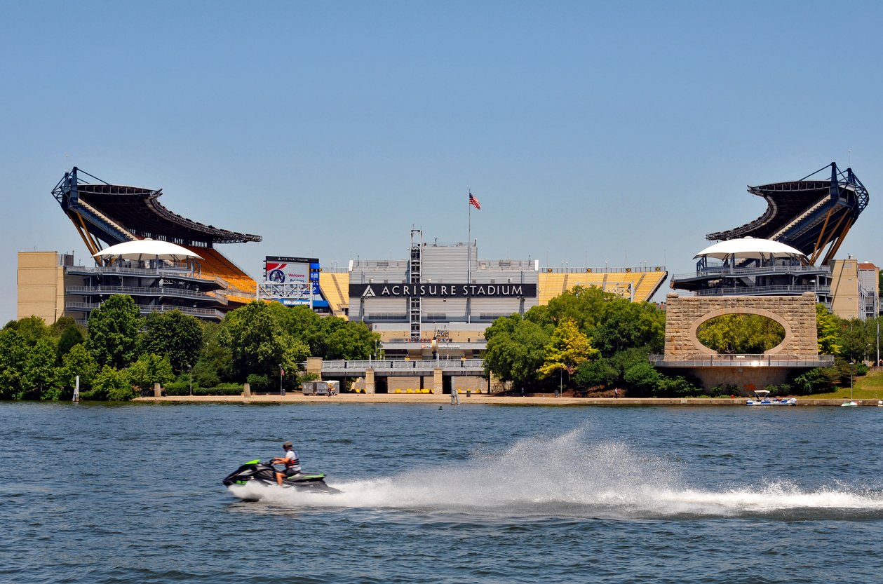 A person rides a jet ski on a river in front of Acrisure Stadium, a large football stadium with distinctive open corners and an arch, on a clear sunny day. Trees line the shore between the water and the stadium.