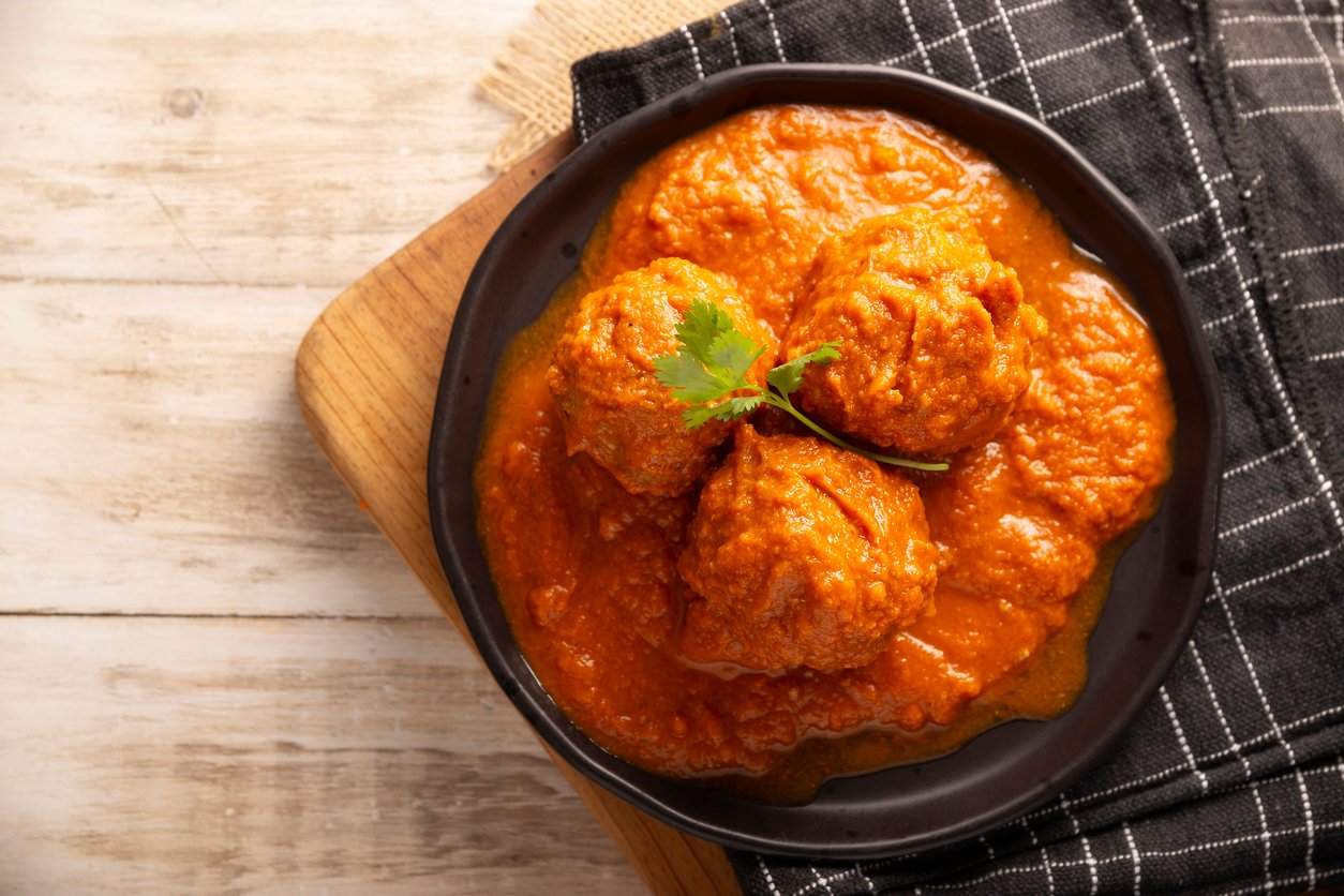 A black plate with three meatballs in rich, orange tomato sauce, garnished with a cilantro sprig, placed on a wooden board beside a black and white checkered cloth.