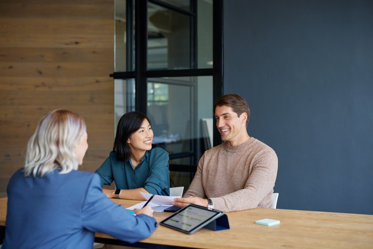 A smiling couple sits at a desk with a business professional, holding documents and having a discussion. A tablet and papers are on the wooden table in a modern office setting.