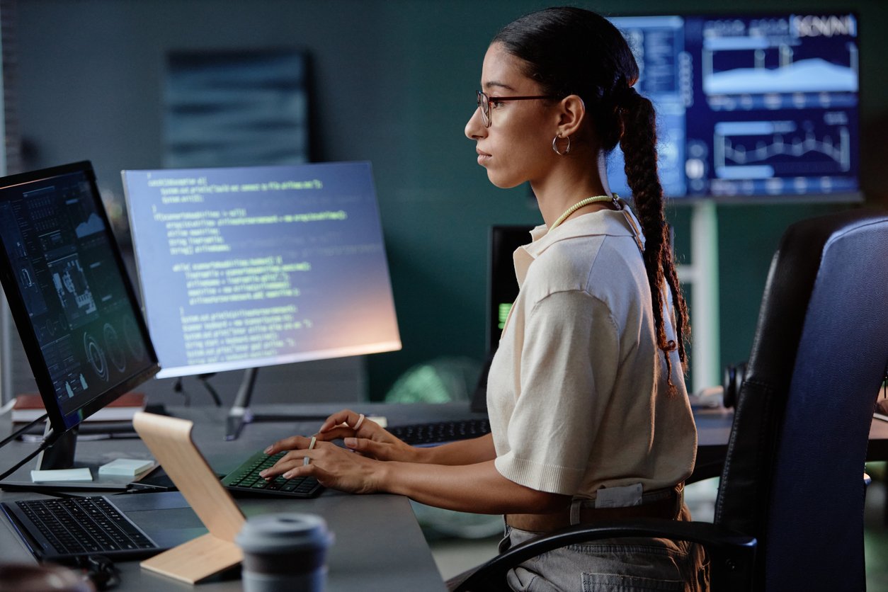 A woman with glasses and a braided ponytail works at a computer with code on the monitor, in a modern office with charts and data displayed on large screens in the background.