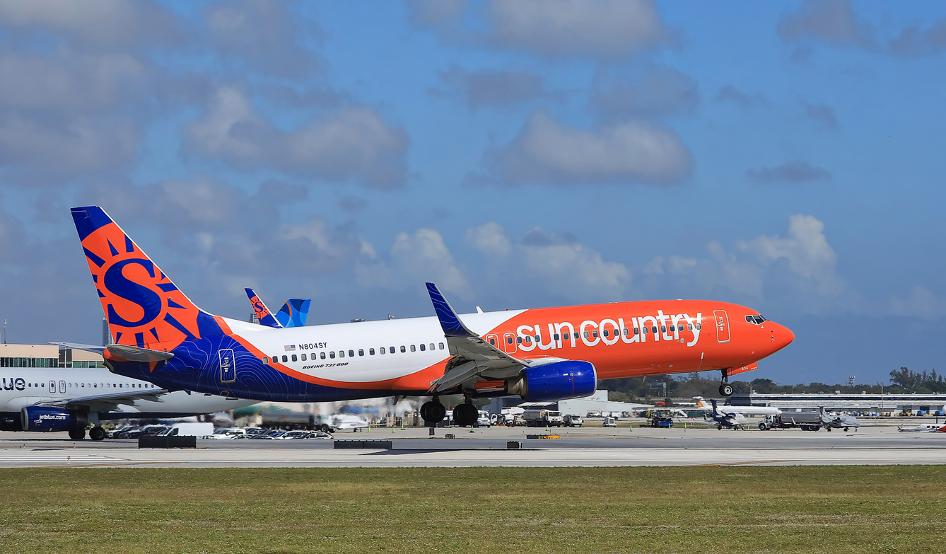 A Sun Country Airlines plane painted orange and blue is taking off from an airport runway on a clear day, with airport buildings and another plane in the background.