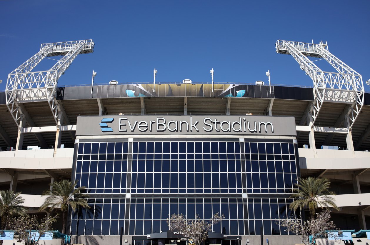 Exterior view of EverBank Stadium, featuring its name on a large sign above glass windows, with white support structures, palm trees, and a clear blue sky in the background.