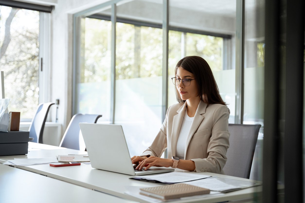 A woman wearing glasses and a beige blazer sits at a desk in a bright office, typing on a laptop. Papers, a notebook, and a phone are on the desk, and large windows let in natural light.