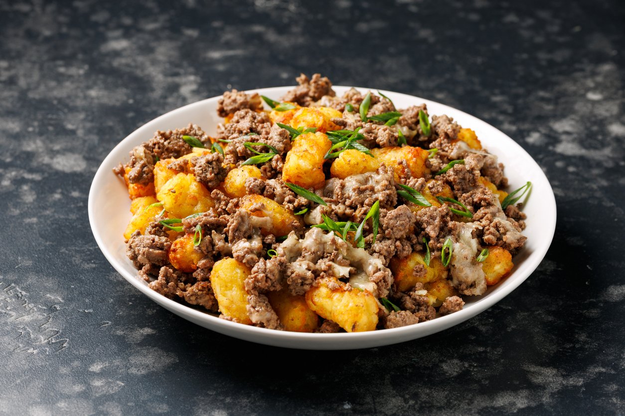 A white bowl filled with crispy tater tots topped with seasoned ground beef, melted cheese, and chopped green onions on a dark, textured surface.