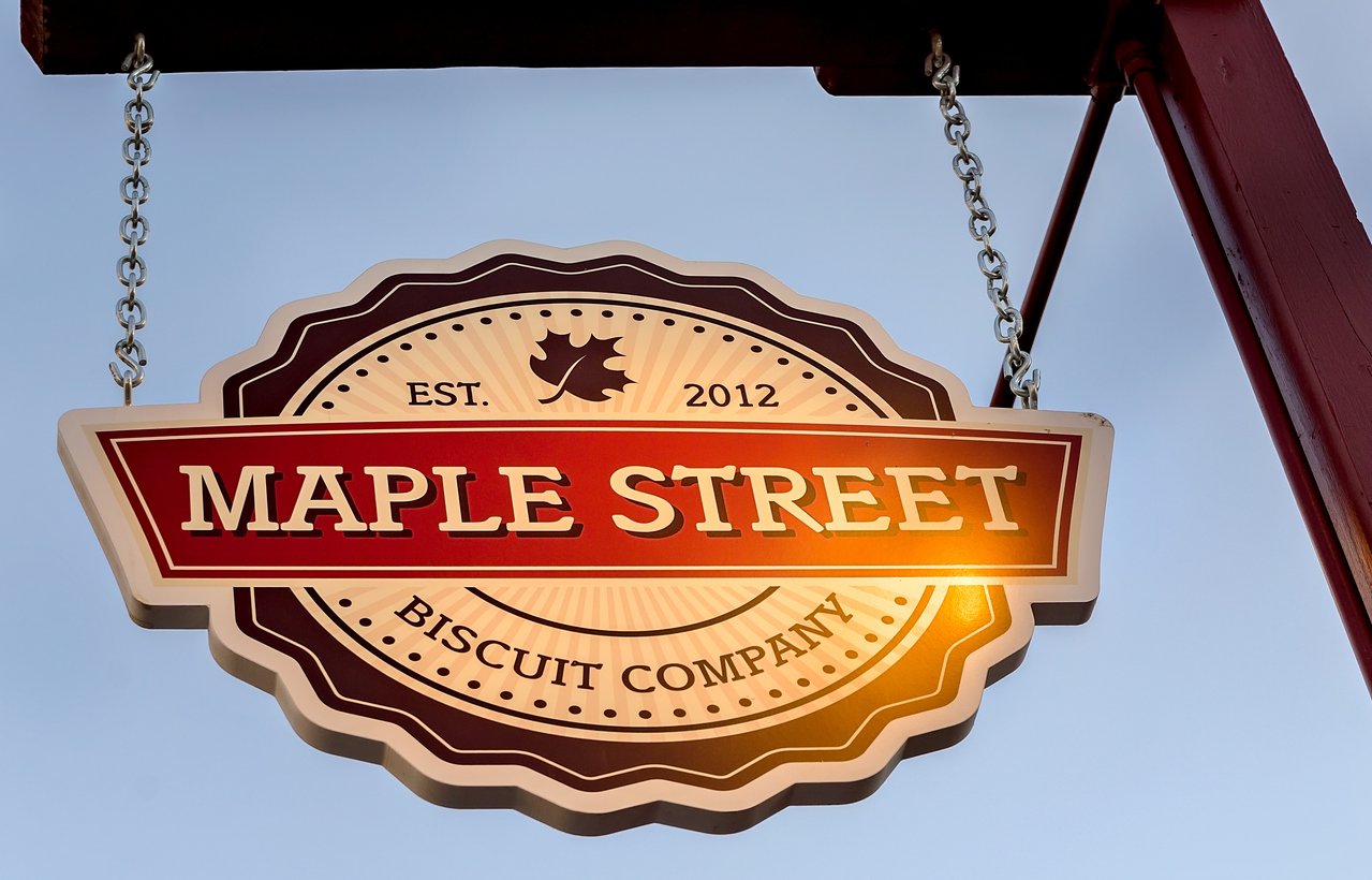 Hanging sign for Maple Street Biscuit Company, featuring a maple leaf, "EST. 2012," and decorative borders, illuminated by sunlight against a clear blue sky.