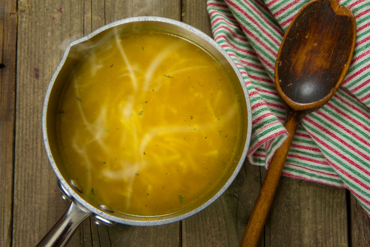 A pot of hot noodle soup sits on a wooden surface next to a striped kitchen towel and a wooden spoon. Steam rises from the soup, which contains thin noodles in a clear broth.