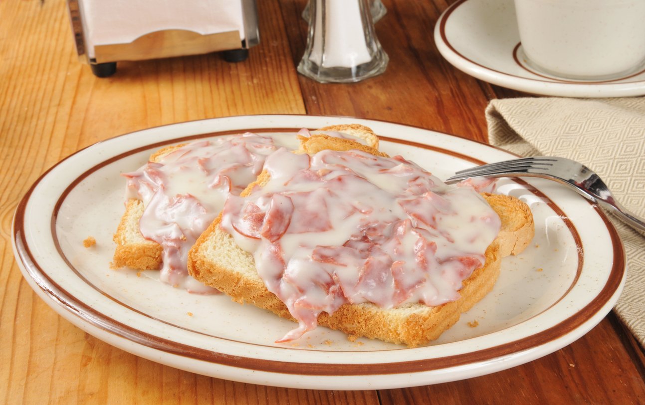 Two slices of toast topped with chipped beef and creamy white gravy are served on a plate with a fork, napkin, and coffee cup on a wooden table.
