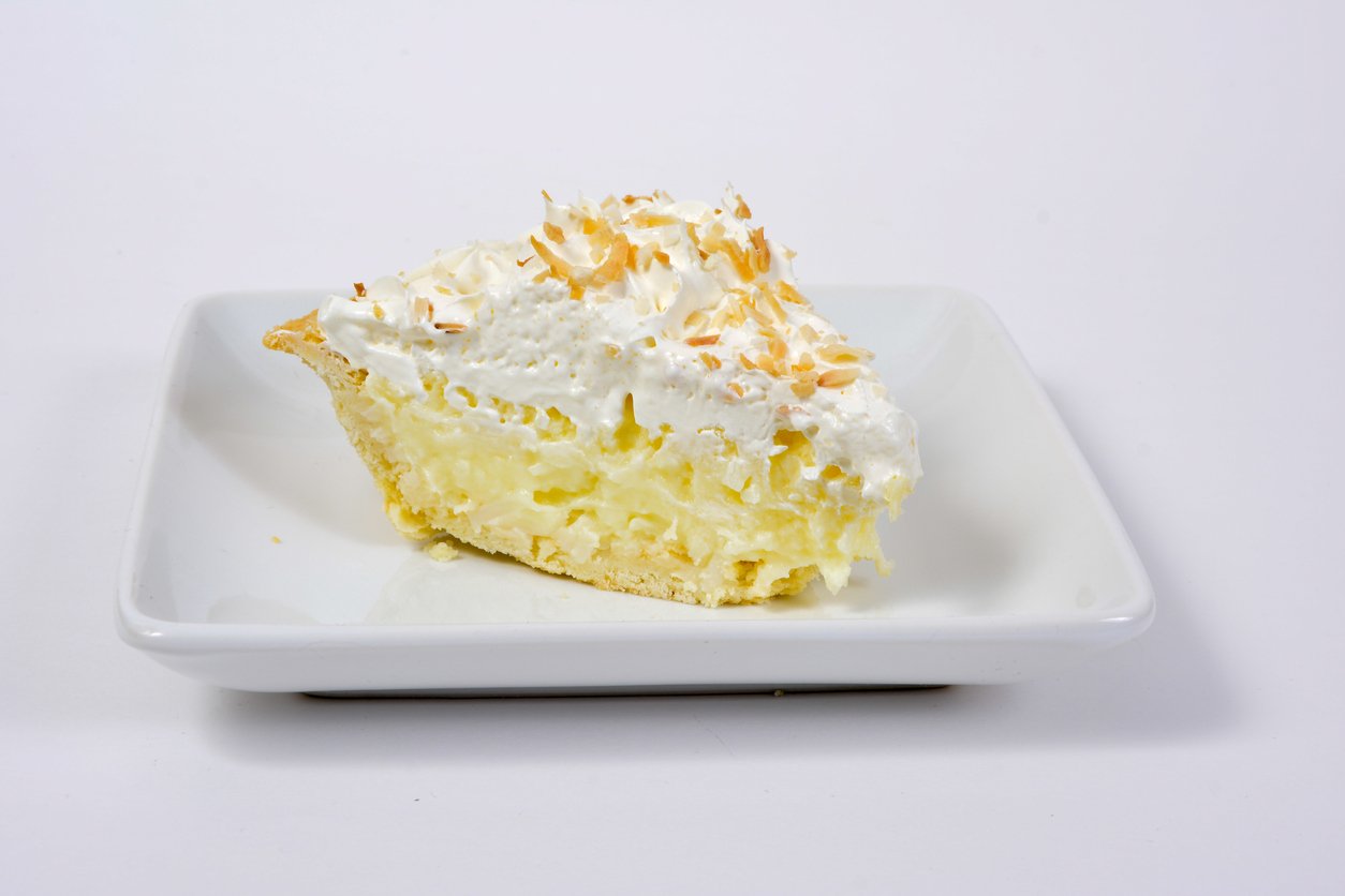 A slice of coconut cream pie topped with whipped cream and toasted coconut flakes sits on a white square plate against a plain white background.