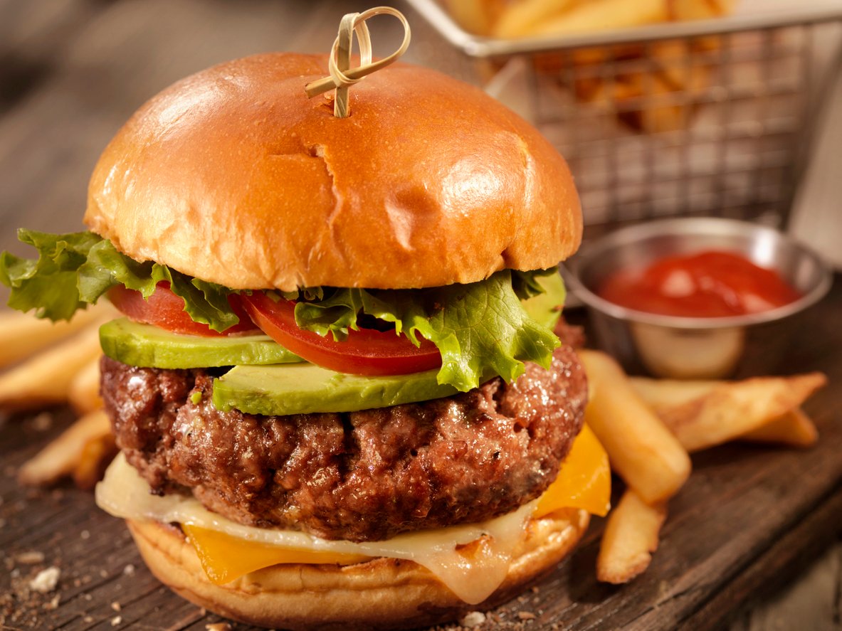 A juicy hamburger with lettuce, tomato, and avocado on a toasted bun, served with melted cheese, crispy fries, and a small metal cup of ketchup in the background.