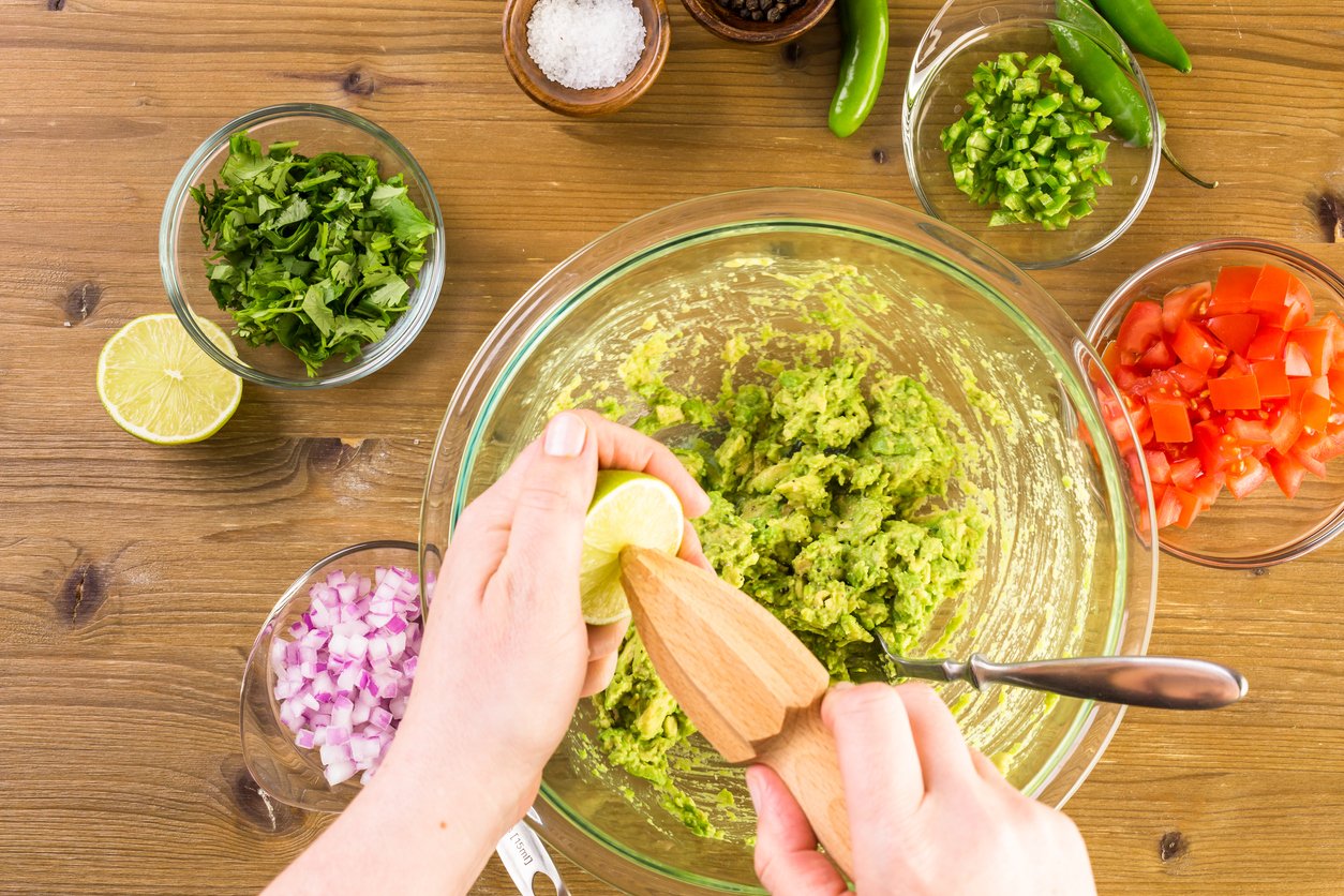A person squeezes a lime into a bowl of mashed avocado, surrounded by bowls of chopped tomato, onion, cilantro, jalapeño, and seasonings on a wooden table.