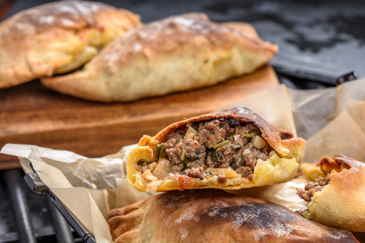 A close-up of baked empanadas, one cut open to show a savory filling of ground meat, onions, and herbs, resting on parchment paper with more empanadas and a wooden board in the background.