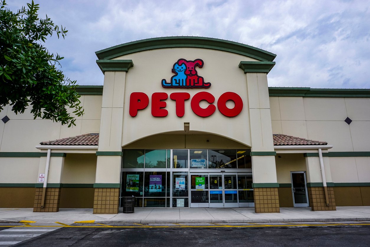 The front entrance of a PETCO store with large red letters and animal logo above glass double doors, surrounded by a beige building and a cloudy sky overhead.