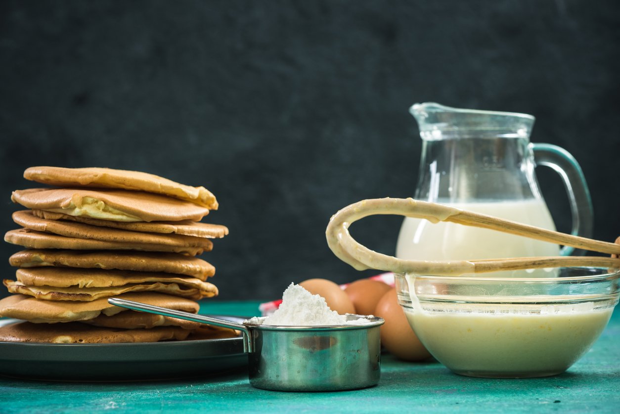 A stack of pancakes sits beside a measuring cup of flour, a bowl of batter with a whisk, eggs, and a glass pitcher of milk, all arranged on a turquoise surface against a dark background.