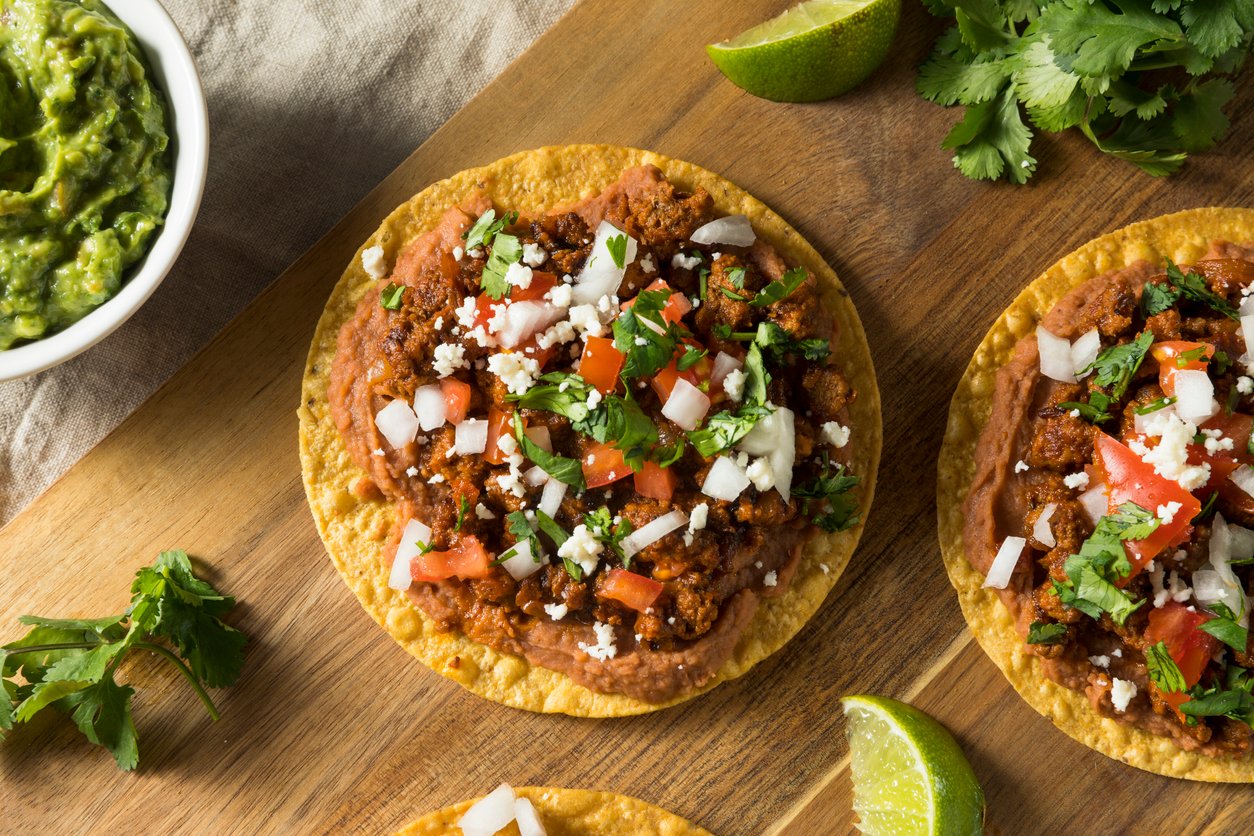 A tostada topped with seasoned ground meat, chopped tomatoes, onions, cilantro, and crumbled cheese sits on a wooden board near lime wedges, fresh cilantro, and a bowl of guacamole.