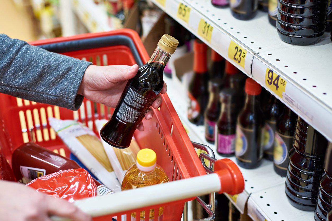 A person places a bottle of soy sauce into a shopping basket filled with groceries, while standing in front of a store shelf stocked with similar bottles and price tags.
