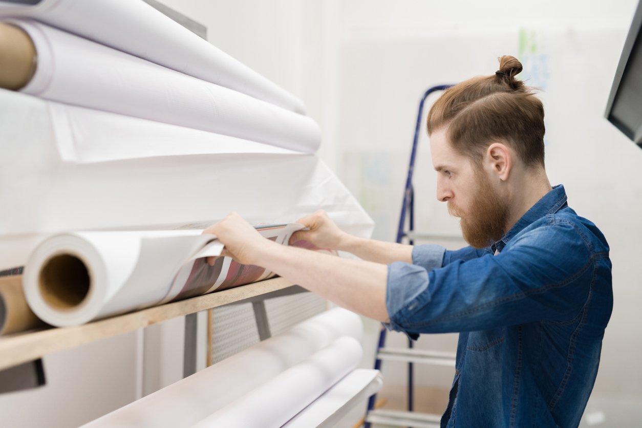 A man with a beard and a top knot, wearing a denim shirt, examines large rolls of paper or prints on a shelf in a bright workspace. A ladder is visible in the background.