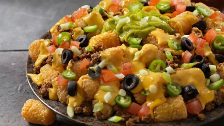 A plate of loaded tater tots topped with melted cheese, ground beef, diced tomatoes, sliced black olives, jalapeños, green onions, and a generous scoop of guacamole.