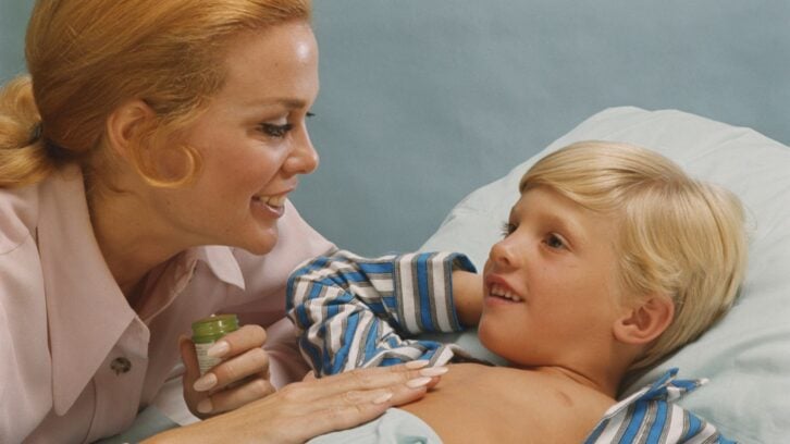 A woman smiles at a young boy lying in bed, holding an open jar of ointment as she applies it to his chest. The boy is wearing blue striped pajamas and looks up at her with a smile.