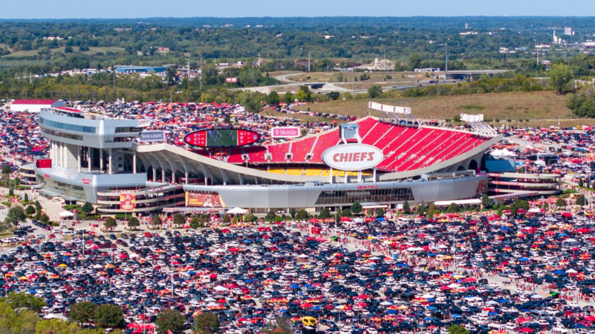 Aerial view of Arrowhead Stadium filled with red seats and a crowded parking lot, with numerous vehicles, on a clear day. The Kansas City Chiefs logo is visible on the stadium.