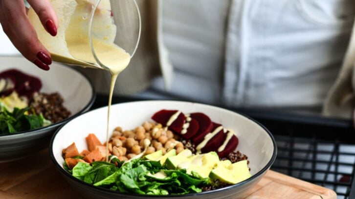 A person pours a creamy dressing over a bowl of salad containing spinach, chickpeas, sweet potatoes, beets, avocado, and quinoa.