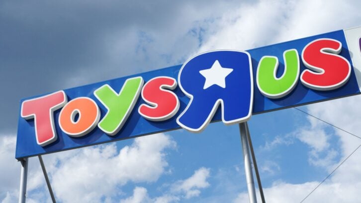A colorful Toys "R" Us store sign with red, green, blue, yellow, and white letters stands against a blue sky with scattered clouds.