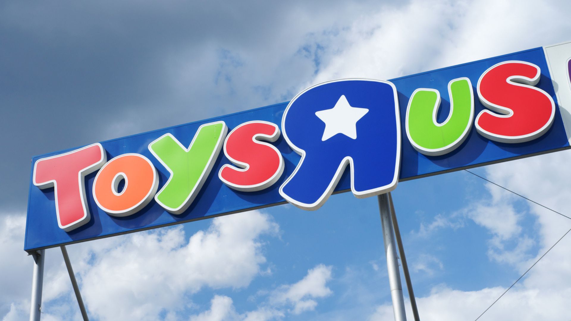 A colorful Toys "R" Us store sign with red, green, blue, yellow, and white letters stands against a blue sky with scattered clouds.
