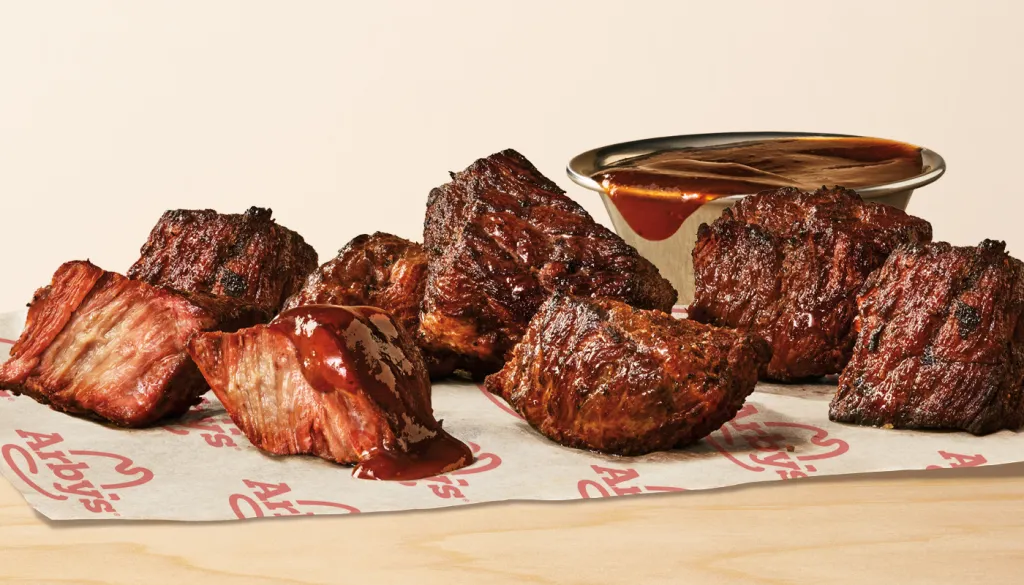 Chunks of smoked beef brisket on Arby’s branded paper, some pieces with barbecue sauce, showcase this new fast-food favorite, with a metal cup of barbecue sauce in the background on a light wood surface.