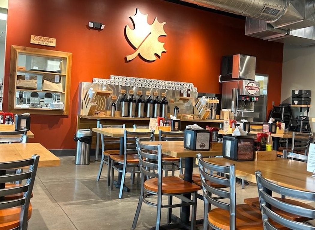 A casual restaurant interior with wooden tables and metal chairs, a drink station along an orange wall with a large leaf logo, condiments on tables, and a modern, clean atmosphere.