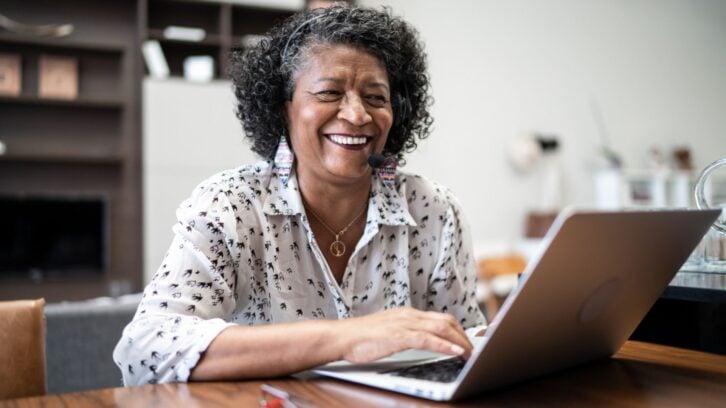 An older woman with curly hair and colorful earrings smiles while using a laptop at a wooden table in a cozy, modern home environment.