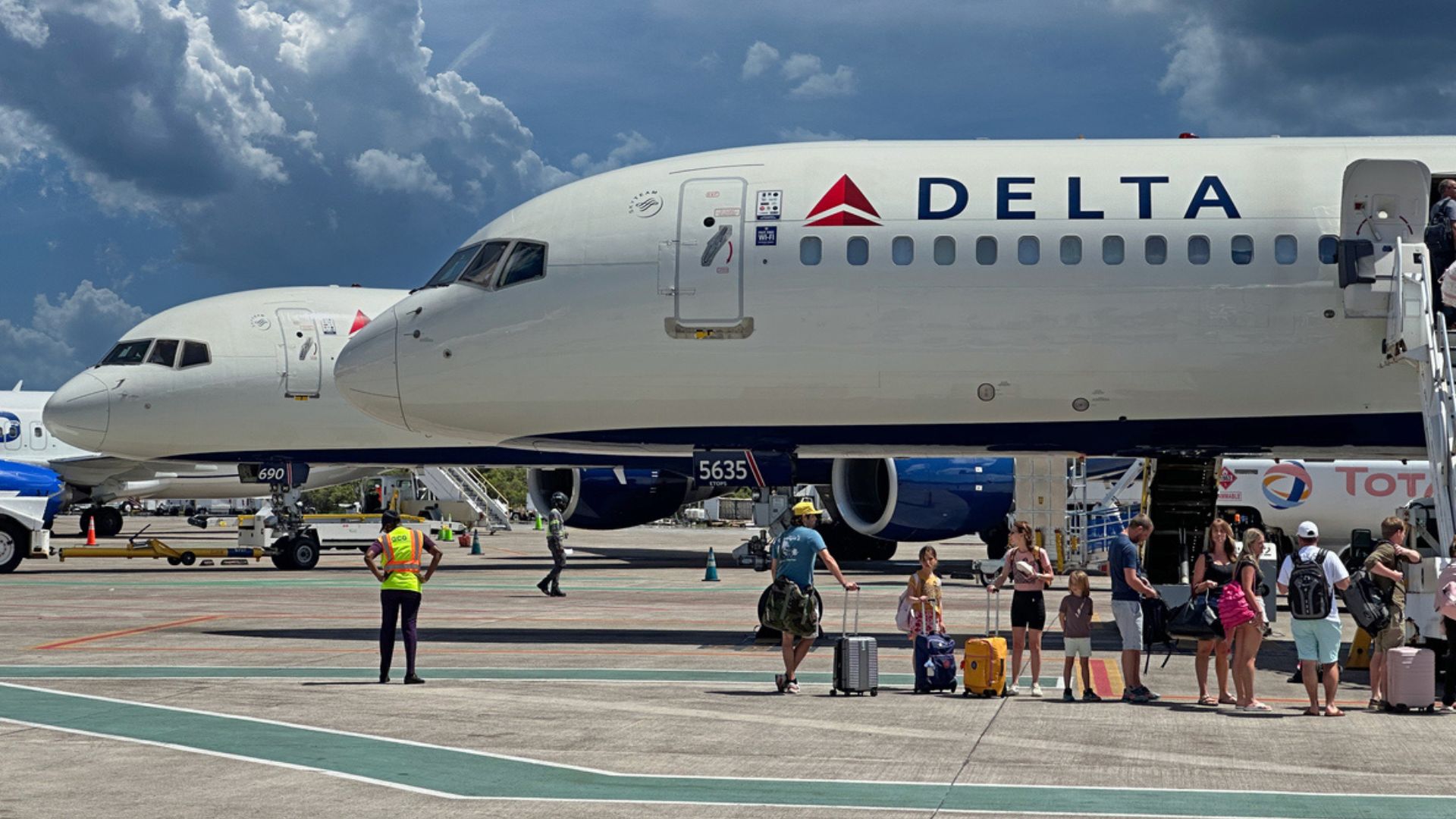 Passengers with luggage board a Delta airplane via stairs on the tarmac, while a worker in a reflective vest stands nearby. Another airplane is visible in the background under a dramatic cloudy sky.