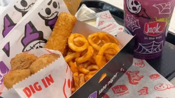Enjoy a new fast-food experience with a Jack in the Box meal featuring curly fries, chicken nuggets, a mozzarella stick, and a purple drink in a Halloween-themed cup, all served on branded paper and a tray.