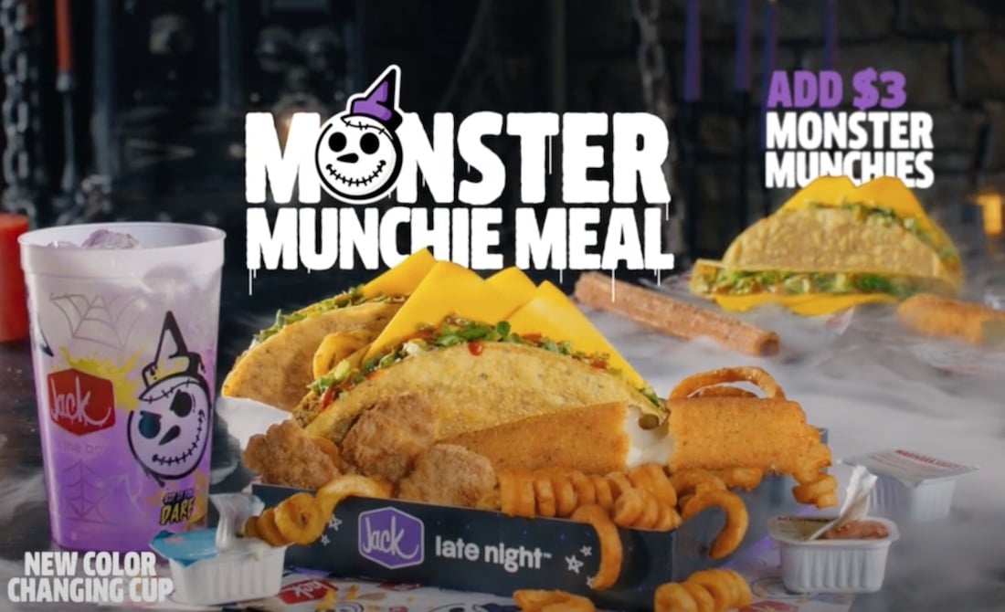 A spooky-themed new fast-food tray displays two tacos, curly fries, mozzarella sticks, chicken strips, and a cheeseburger with Halloween graphics. A purple color-changing drink cup sits on the side. Text advertises the "Monster Munchie Meal.