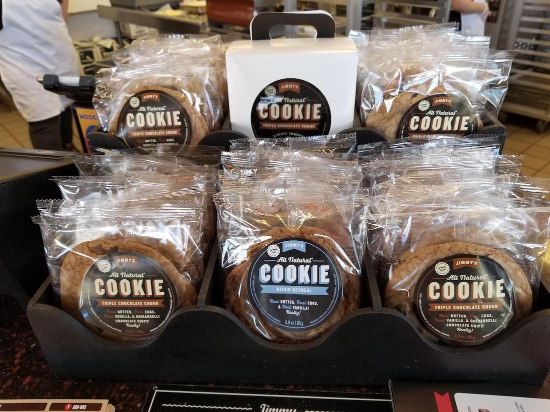 Packets of "Triple Chocolate Chunk" cookies are displayed in a black stand at a counter, with a bakery or café setting visible in the background. Each cookie is individually wrapped in plastic.