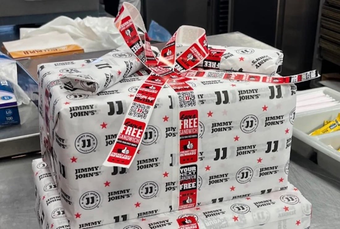 Two stacked boxes wrapped in Jimmy John’s branded sandwich paper and Jimmy John’s promotional tape, forming a bow on top. The packages sit on a countertop in a commercial kitchen.