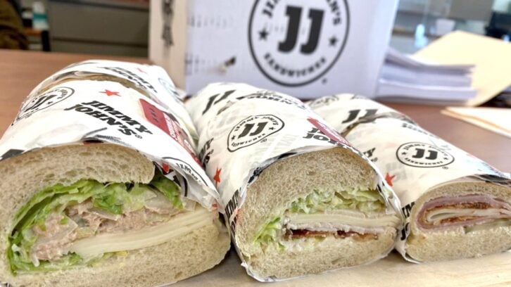 Three wrapped sub sandwiches from Jimmy John’s are cut open to show their fillings, which include lettuce, sliced meats, and cheese. A Jimmy John’s box and papers are visible in the background.