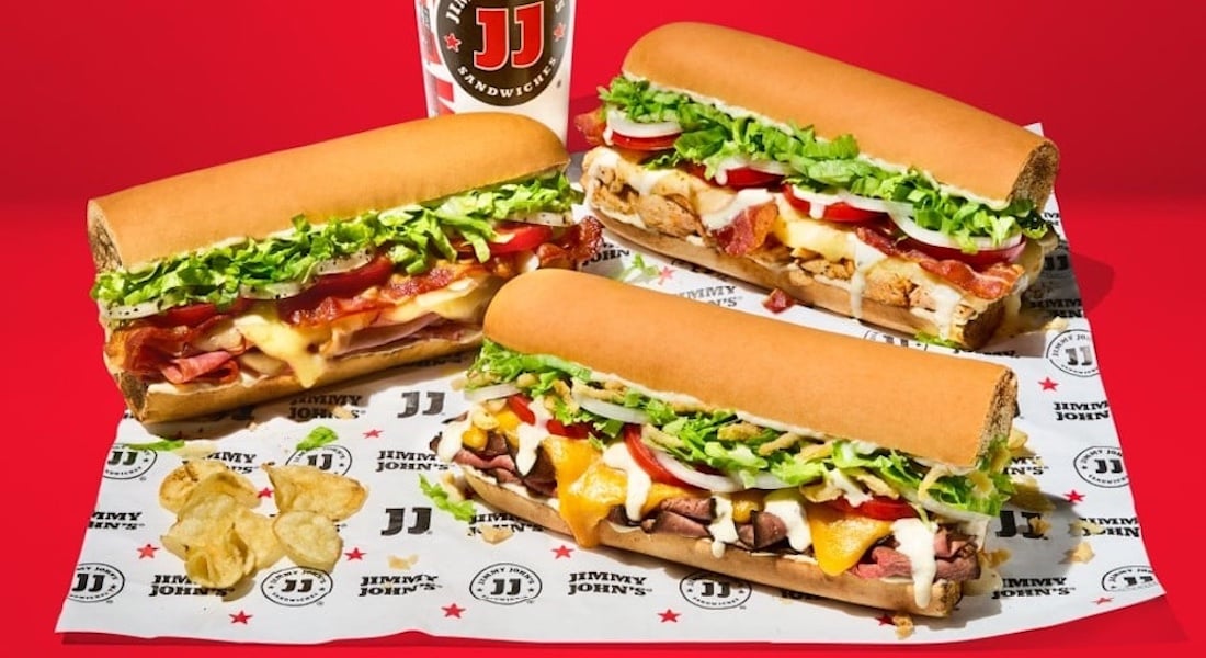 Three large sub sandwiches filled with meats, cheese, lettuce, tomato, and onions are displayed on Jimmy John’s branded paper, with a few potato chips and a soft drink cup in the background, set against a bright red, new fast-food backdrop.
