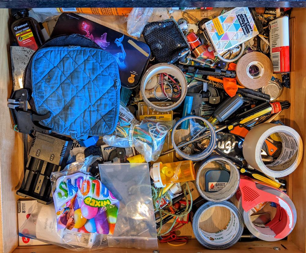 A cluttered drawer filled with assorted items, including tape rolls, scissors, pens, batteries, a blue oven mitt, balloons, rubber bands, cables, and various small tools and office supplies.