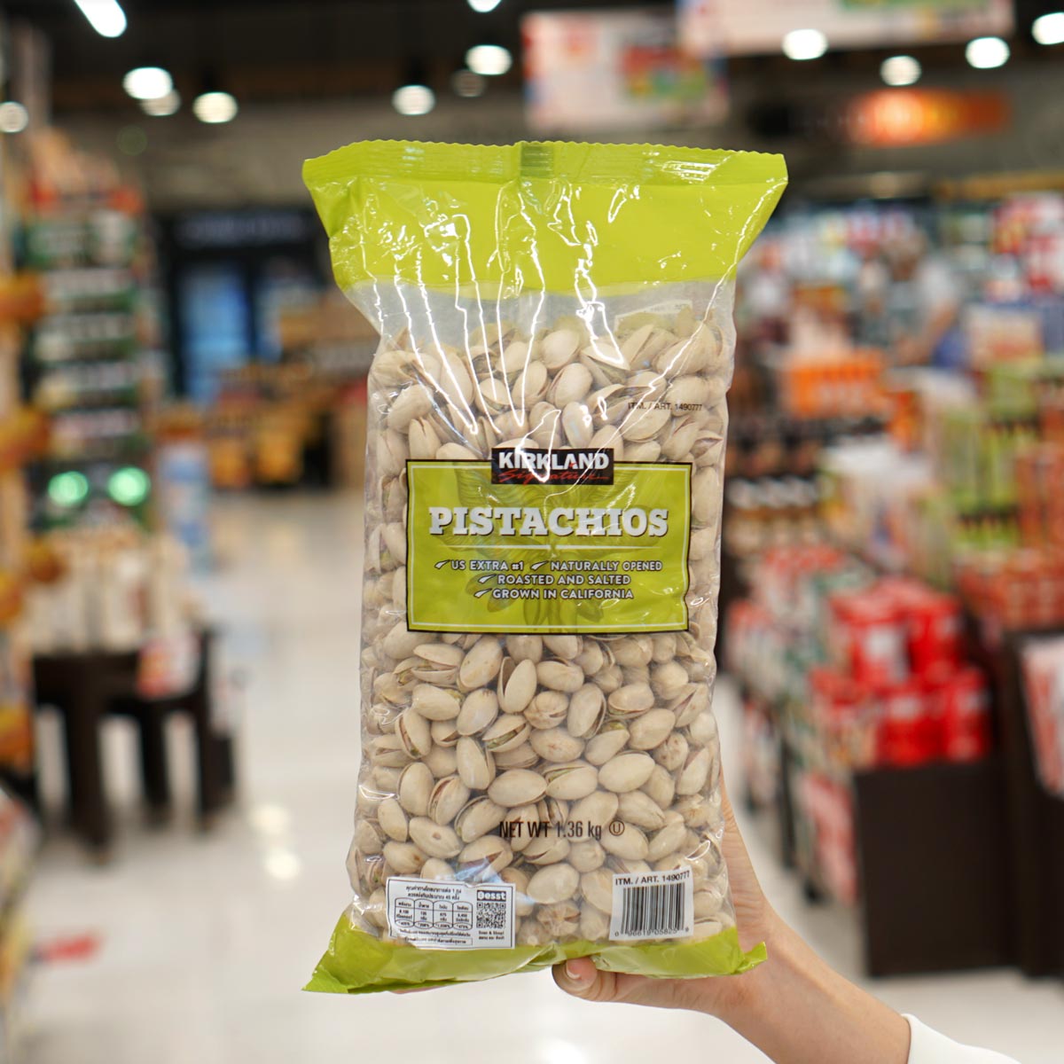A hand holds a large, clear bag of Kirkland brand pistachios—one of the Costco products loved by shoppers—in a grocery store aisle lined with fully stocked shelves.