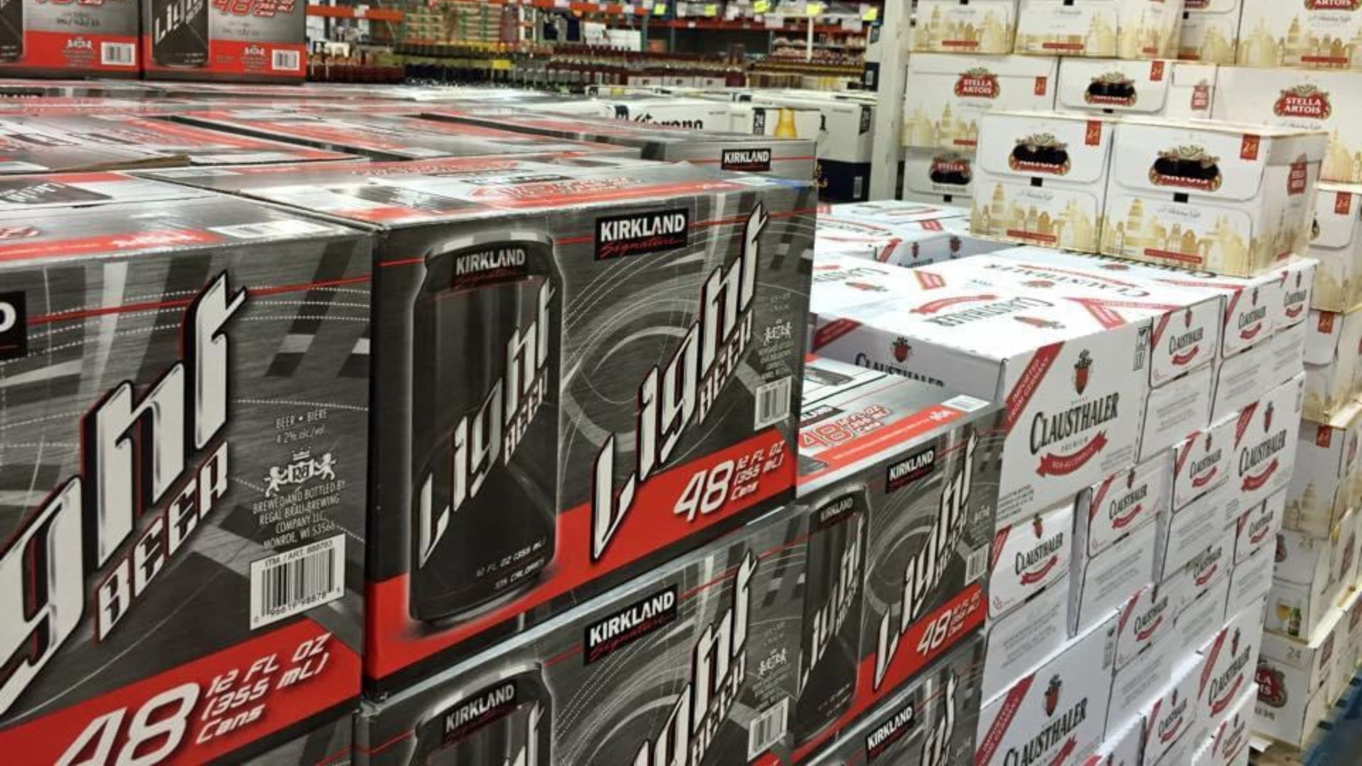 Stacks of beer cases, including Kirkland light beer in red and gray boxes and Clausthaler in white boxes, are displayed on shelves in a warehouse-style store.