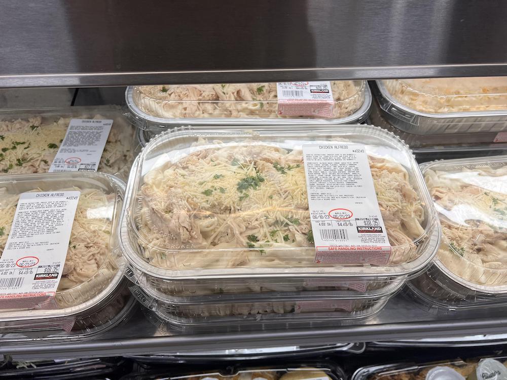 Plastic containers filled with uncooked pasta topped with shredded cheese and herbs are stacked on a refrigerated grocery store shelf, each with a white and red nutrition label.