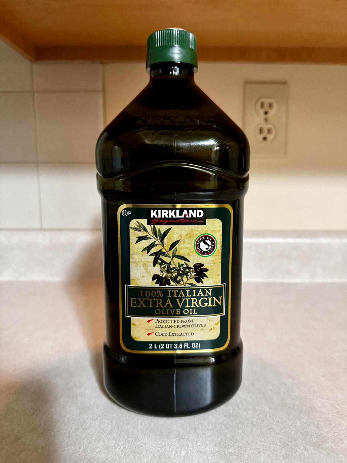 A large 2-liter bottle of Kirkland 100% Italian Extra Virgin Olive Oil, one of the Costco products loved by shoppers, sits on a kitchen countertop with a backsplash, electrical outlet, and wooden cabinet in the background.