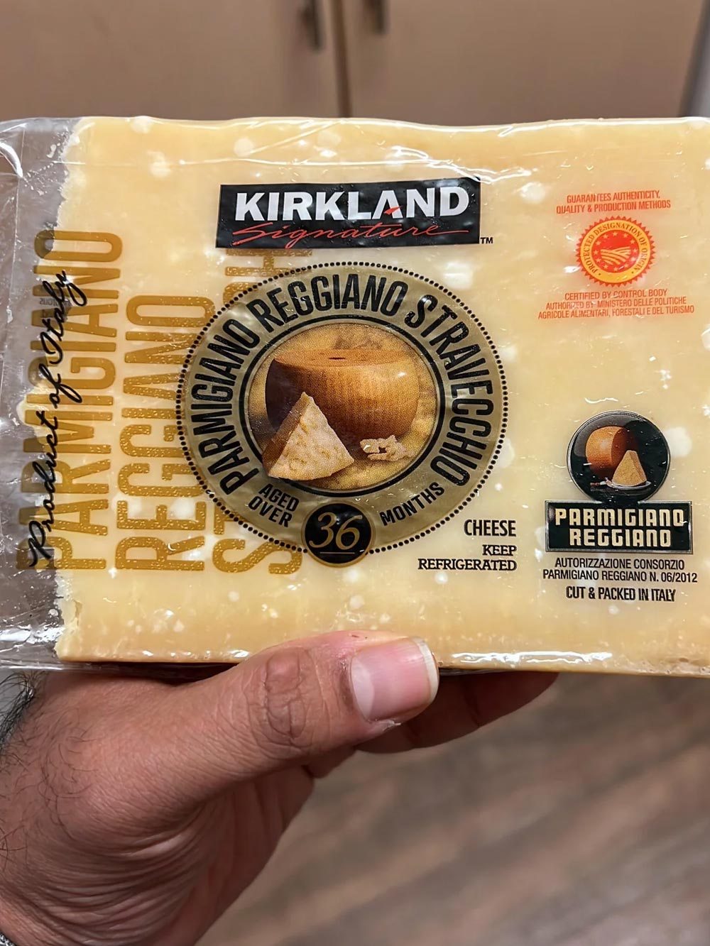 A hand holds a block of Kirkland Signature Parmigiano Reggiano cheese, aged 36 months, in its packaging—a favorite among Costco products loved by shoppers. The label displays a cheese wheel and wedge, Italian certification marks, and storage instructions.