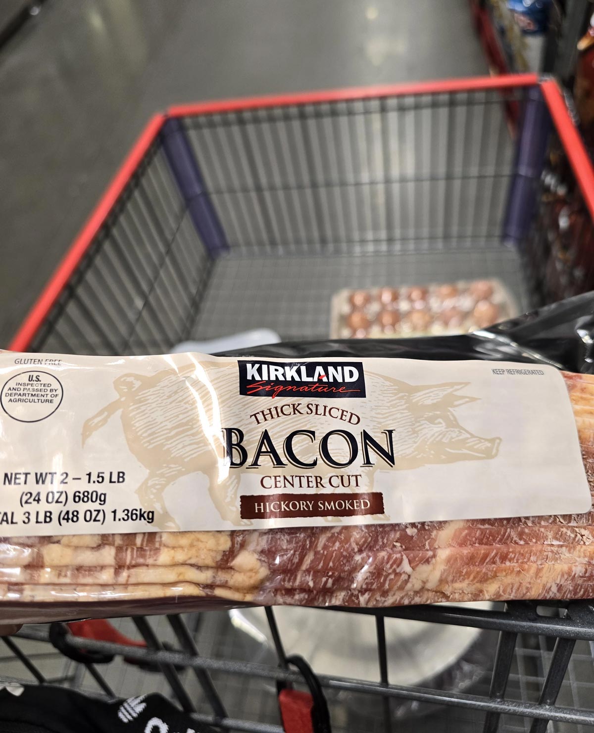 A close-up of a package of Kirkland Signature Thick Sliced Center Cut Hickory Smoked Bacon, one of the Costco products loved by shoppers, in a shopping cart at a store. The package contains 3 pounds (48 oz) of bacon.