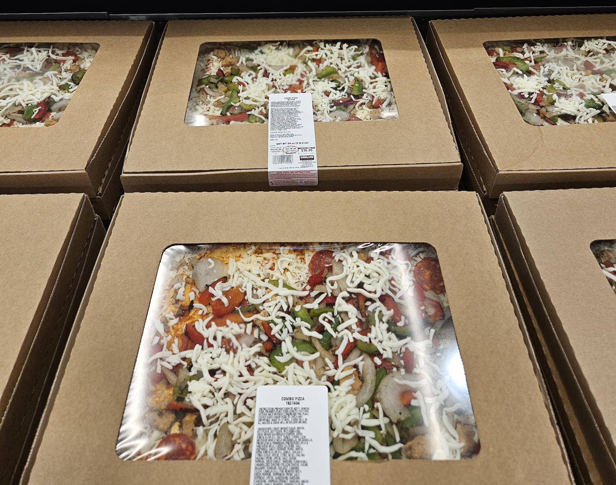 Several large rectangular pizza boxes with clear plastic windows show pizzas topped with cheese, green peppers, onions, and pepperoni, stacked in a display. Each box has a white label with product information.