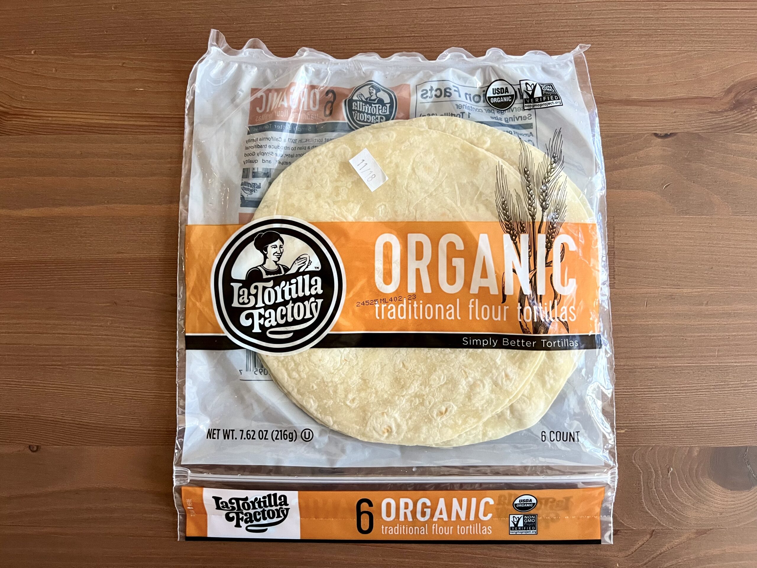A package of La Tortilla Factory organic traditional flour tortillas sits on a wooden table. The clear packaging displays six tortillas and features orange and white branding with a logo and wheat illustration.