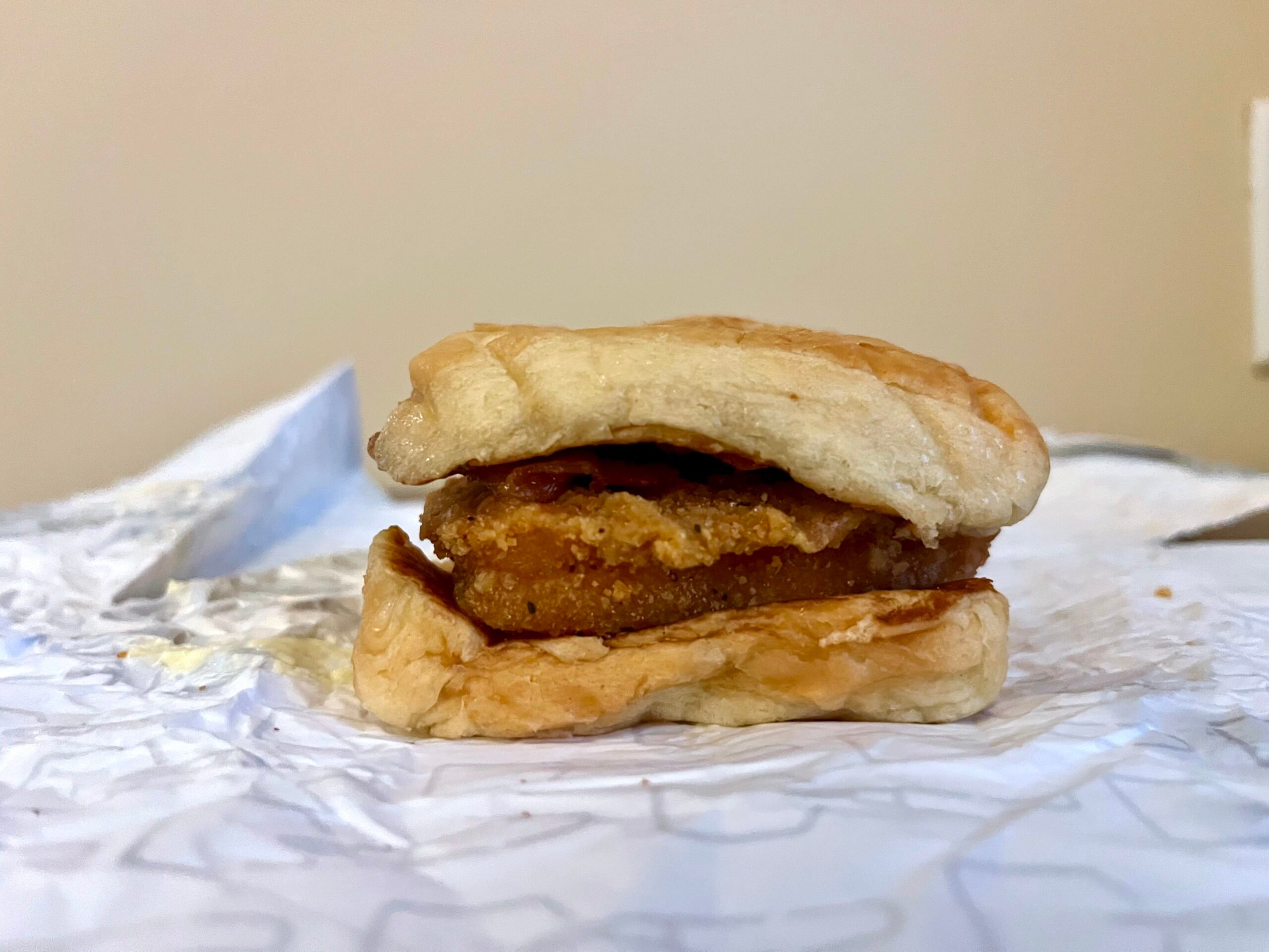 A close-up of a chicken sandwich with a fried chicken patty between two wrinkled, uneven sandwich buns, placed on crumpled white paper with a plain beige background.
