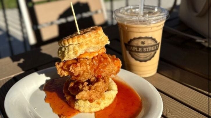 A plate with a stacked biscuit sandwich filled with fried chicken and spicy sauce, secured with a toothpick, sits next to an iced coffee in a clear cup labeled "Maple Street" on an outdoor table.