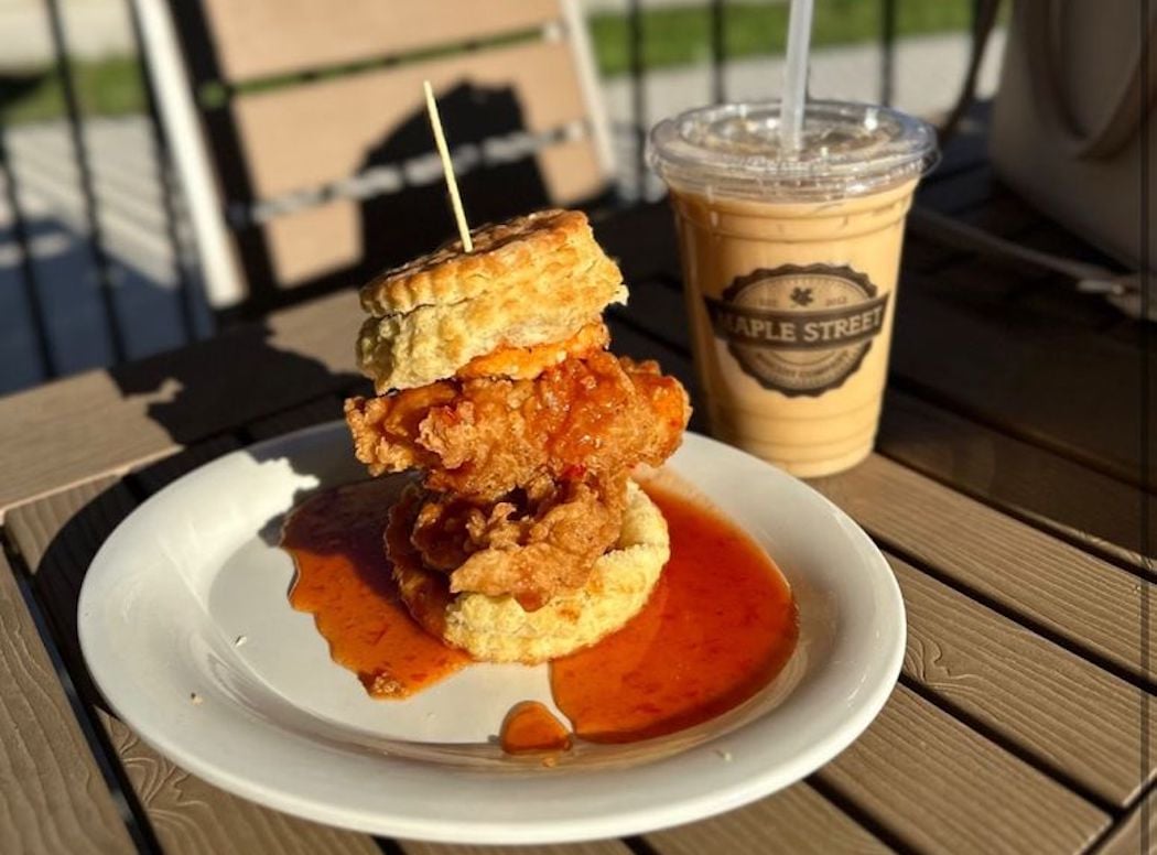 A plate with a biscuit sandwich filled with fried chicken and hot sauce, held together by a toothpick, sits on an outdoor table next to an iced coffee with a “Maple Street” logo. Sunlight casts strong shadows.