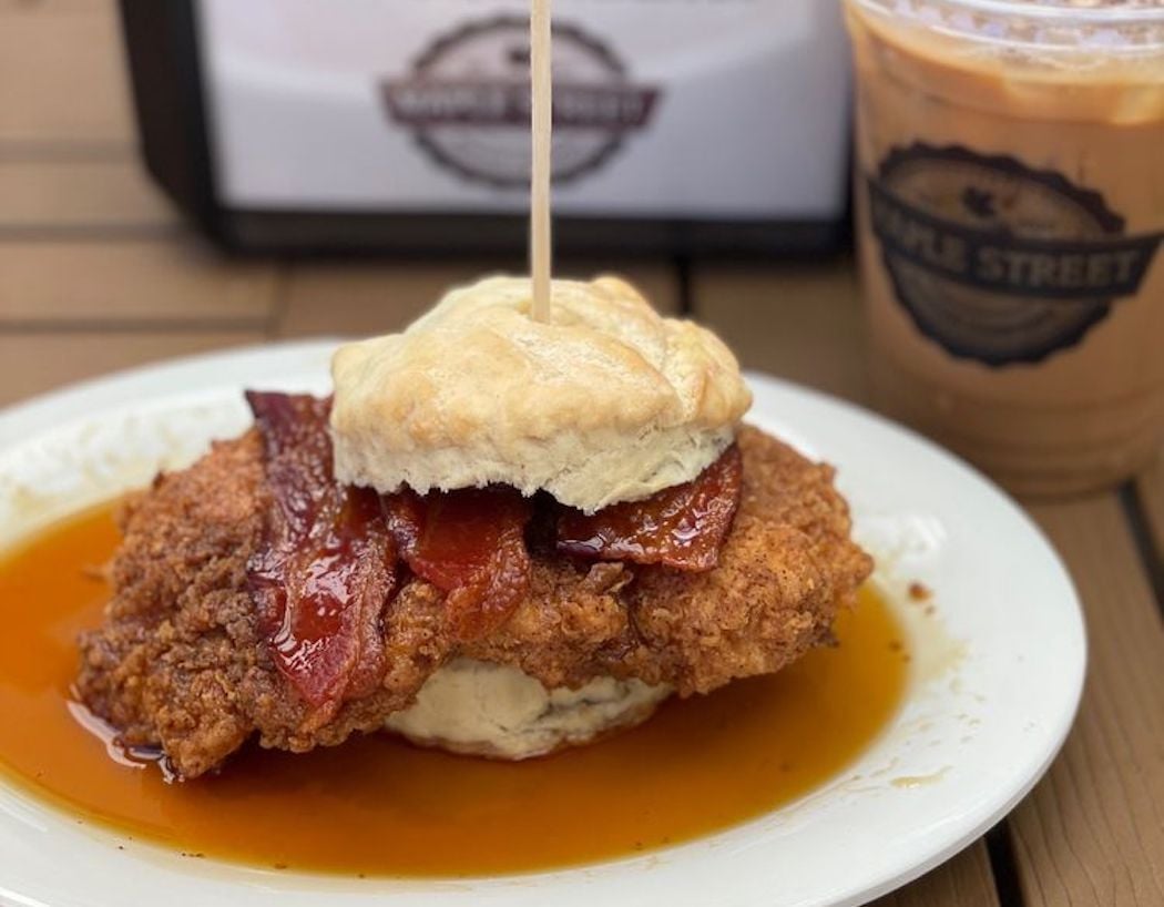 A biscuit sandwich with fried chicken and bacon sits on a white plate covered in syrup, next to a cup of iced coffee on a wooden table.