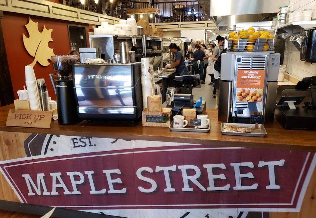 A busy café counter with coffee machines, cups, and pastries, featuring a large "MAPLE STREET" sign and a wooden maple leaf decoration on the wall. Staff are preparing food and drinks in the background.